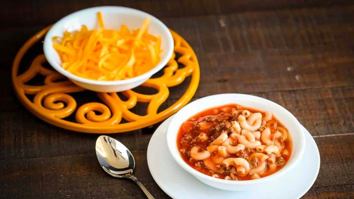 A bowl of macaroni pasta in a tomato and meat sauce sits on a white plate beside a spoon. In the background, there's a bowl of shredded cheese on a decorative yellow tray. All items are placed on a wooden surface.