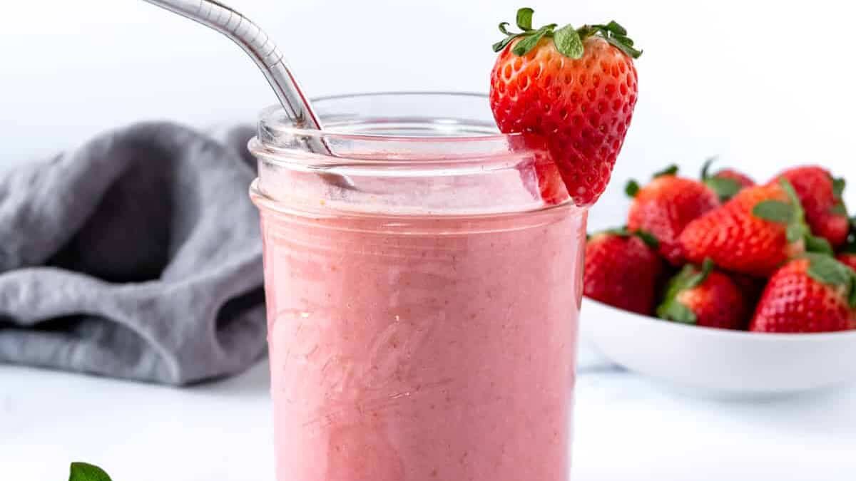A pink smoothie in a glass jar with a metal straw and a fresh strawberry on the rim. A bowl of strawberries is in the background, on a white surface with a gray cloth draped to the side.