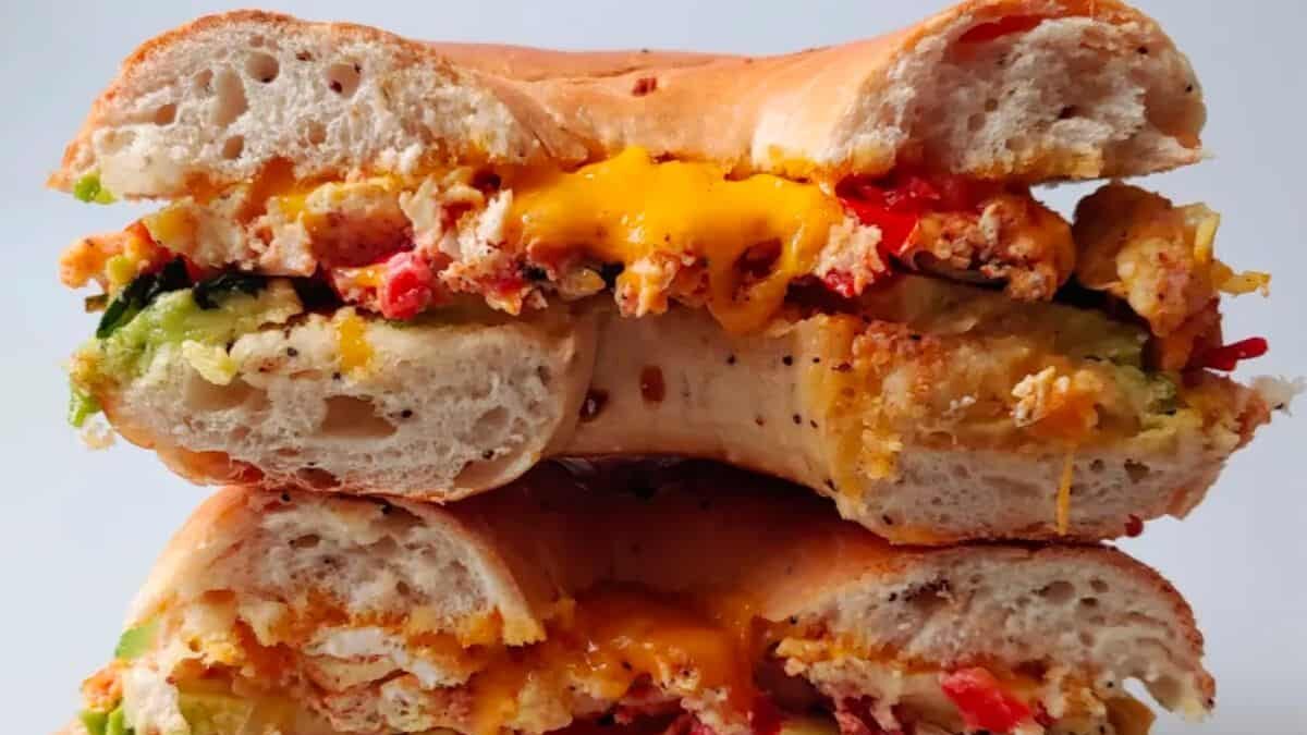 A stacked bagel sandwich is filled with scrambled eggs, melted cheddar cheese, avocado slices, and red bell peppers. The bagel halves are golden brown and slightly toasted, with the filling visibly spilling out, creating an appetizing and colorful display.