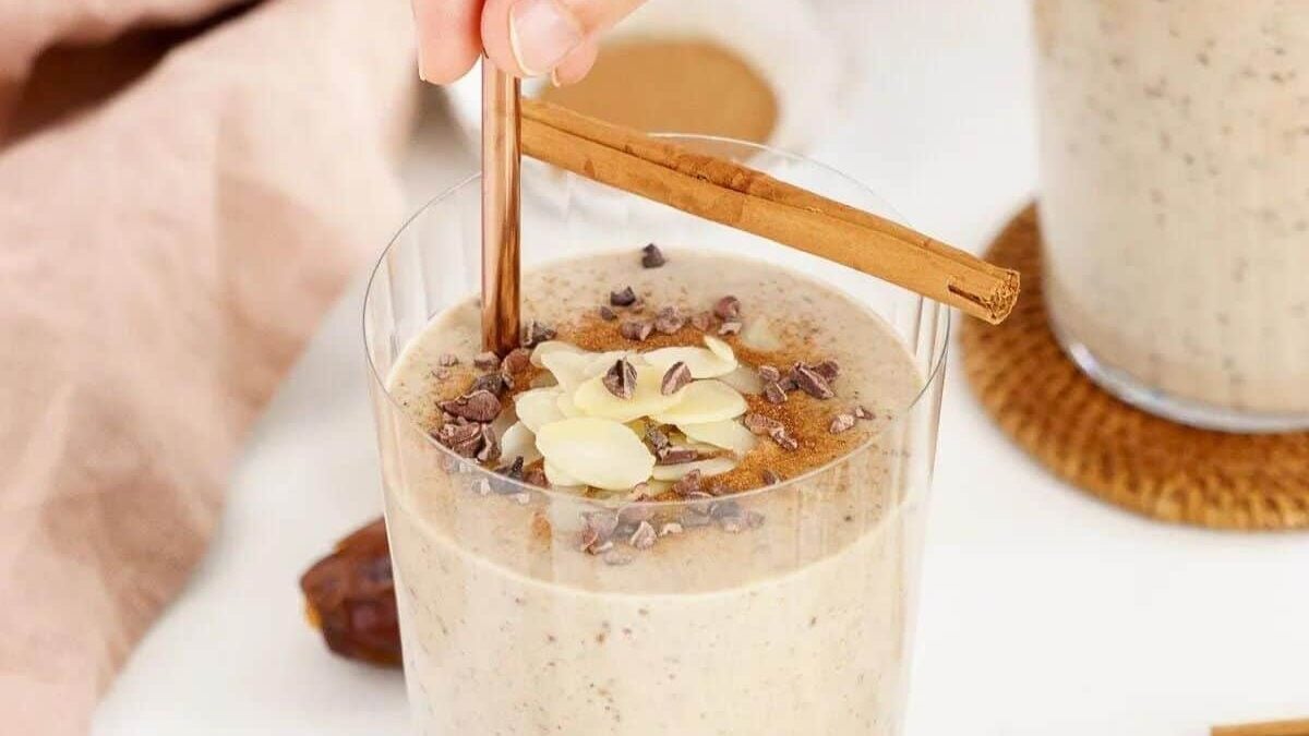 A hand holds a straw stirring a creamy smoothie topped with almond slices and cacao nibs in a glass, garnished with a cinnamon stick. A date lies beside it on a wooden coaster, with a soft beige cloth in the background.