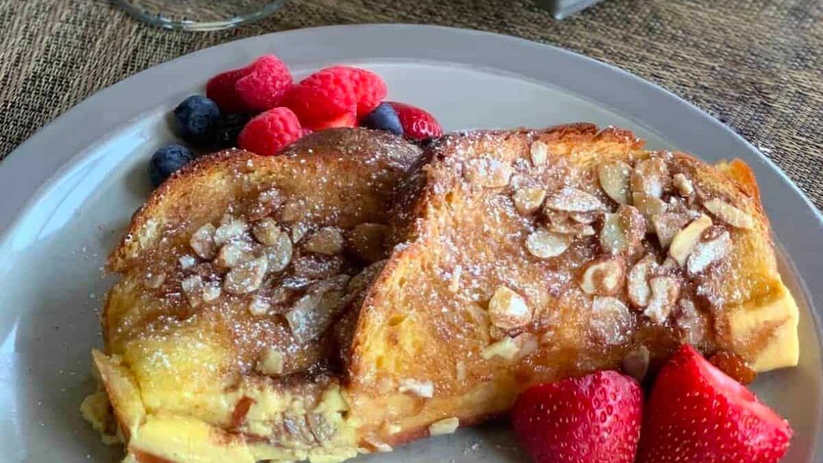A plate of French toast topped with sliced almonds and powdered sugar is served alongside fresh strawberries, raspberries, and blueberries. A detail of a stemmed glass is visible in the background on a textured surface.