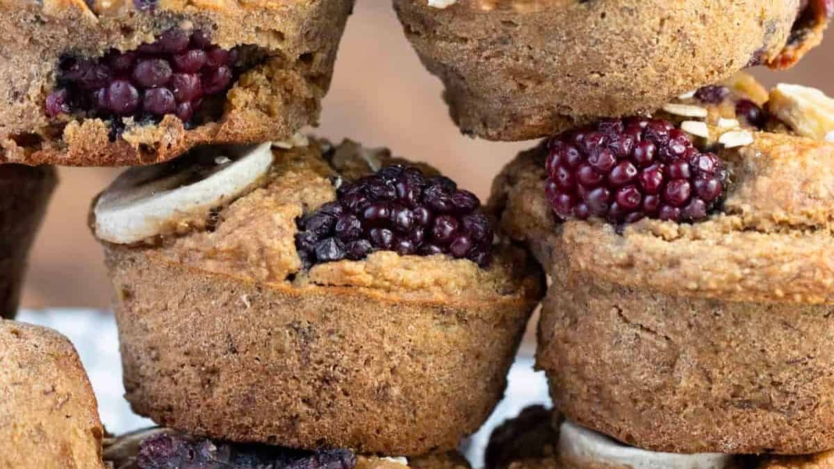 Close-up of stacked muffins with visible blackberries and banana slices baked into them, displaying a rustic, homemade appearance. The muffins have a textured, golden-brown crust.