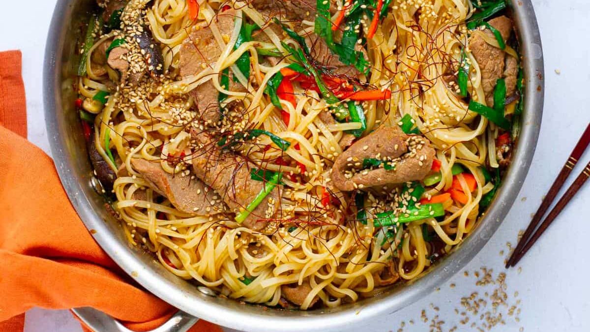 A pan filled with stir-fried noodles, sliced beef, and colorful vegetables like red peppers and green onions. Sesame seeds are sprinkled on top, and the dish is garnished with red pepper strands. A pair of chopsticks and an orange napkin are beside the pan.