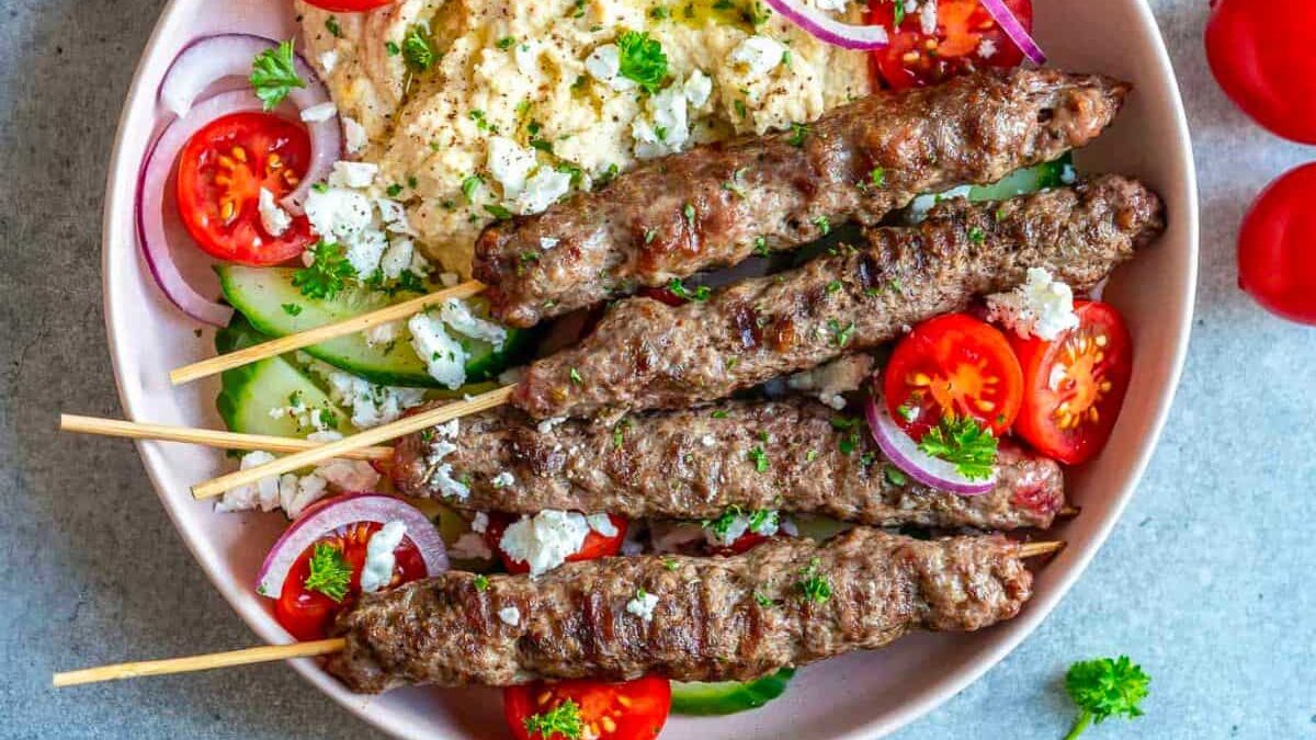 A bowl with skewered grilled meat atop a medley of sliced cucumbers, cherry tomatoes, red onions, and crumbled cheese. Soft hummus is also present, all garnished with chopped parsley on a gray background.