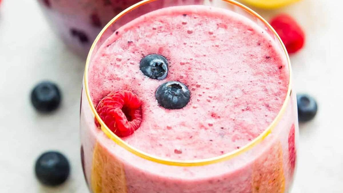 A close-up of a glass filled with a thick pink berry smoothie, topped with two blueberries and a raspberry, with more berries scattered around on a light surface.
