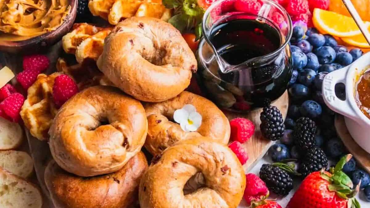 A vibrant breakfast spread featuring a stack of bagels, a small glass jar of syrup, waffles, fresh berries, orange slices, and a bowl of peanut butter, all arranged on a wooden board.