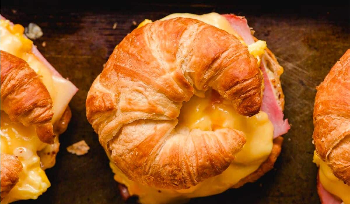 A croissant sandwich with melted cheese, scrambled eggs, and slices of ham. The sandwich is placed on a dark surface, and the croissant's flaky texture is visible. Two similar sandwiches are partially visible in the background.