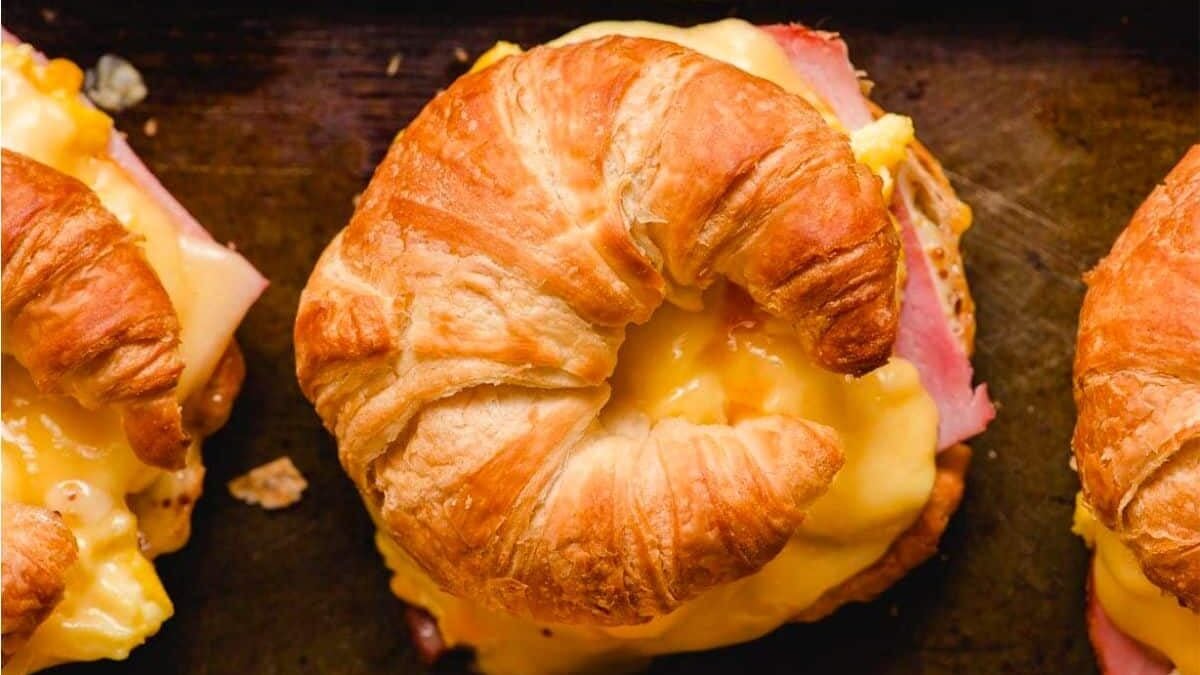 A croissant sandwich with melted cheese, scrambled eggs, and slices of ham. The sandwich is placed on a dark surface, and the croissant's flaky texture is visible. Two similar sandwiches are partially visible in the background.