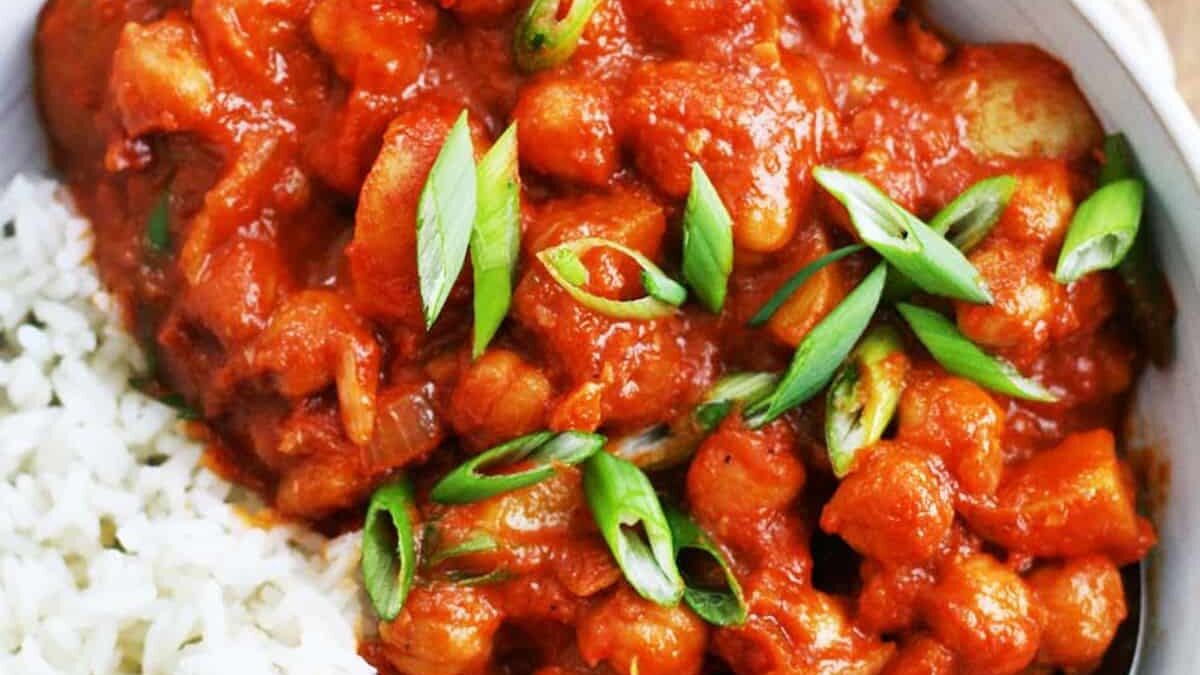 A bowl of white rice and chickpea curry garnished with sliced green onions. The curry features a rich tomato-based sauce, with pieces of chickpeas and potatoes visible throughout.