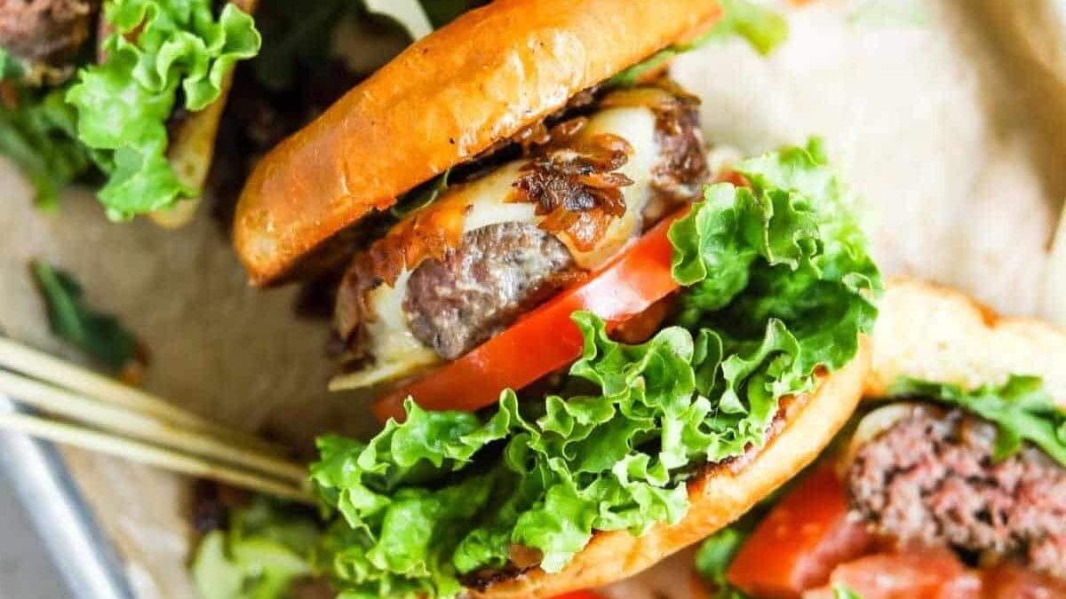 A close-up of a juicy hamburger with a toasted bun, featuring a beef patty, melted cheese, fresh lettuce, a slice of tomato, and caramelized onions. The burger is skewered with a bamboo stick and set on parchment paper.