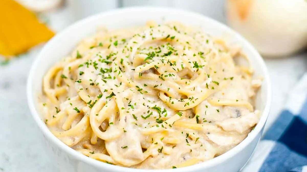 A bowl of creamy pasta topped with freshly chopped herbs. The pasta is coated in a white sauce, and the dish is placed on a table near ingredients like an onion and cheese. A blue and white checkered napkin is partially visible.