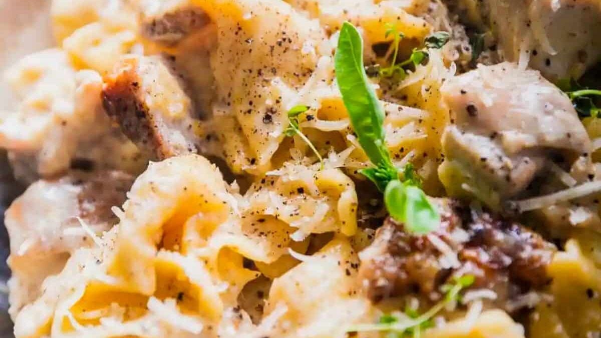 A close-up of a creamy pasta dish featuring tender pieces of chicken, garnished with fresh herbs and sprinkled with grated cheese and black pepper. The pasta appears to be coated in a rich, savory sauce. A single small green basil leaf stands out on top.