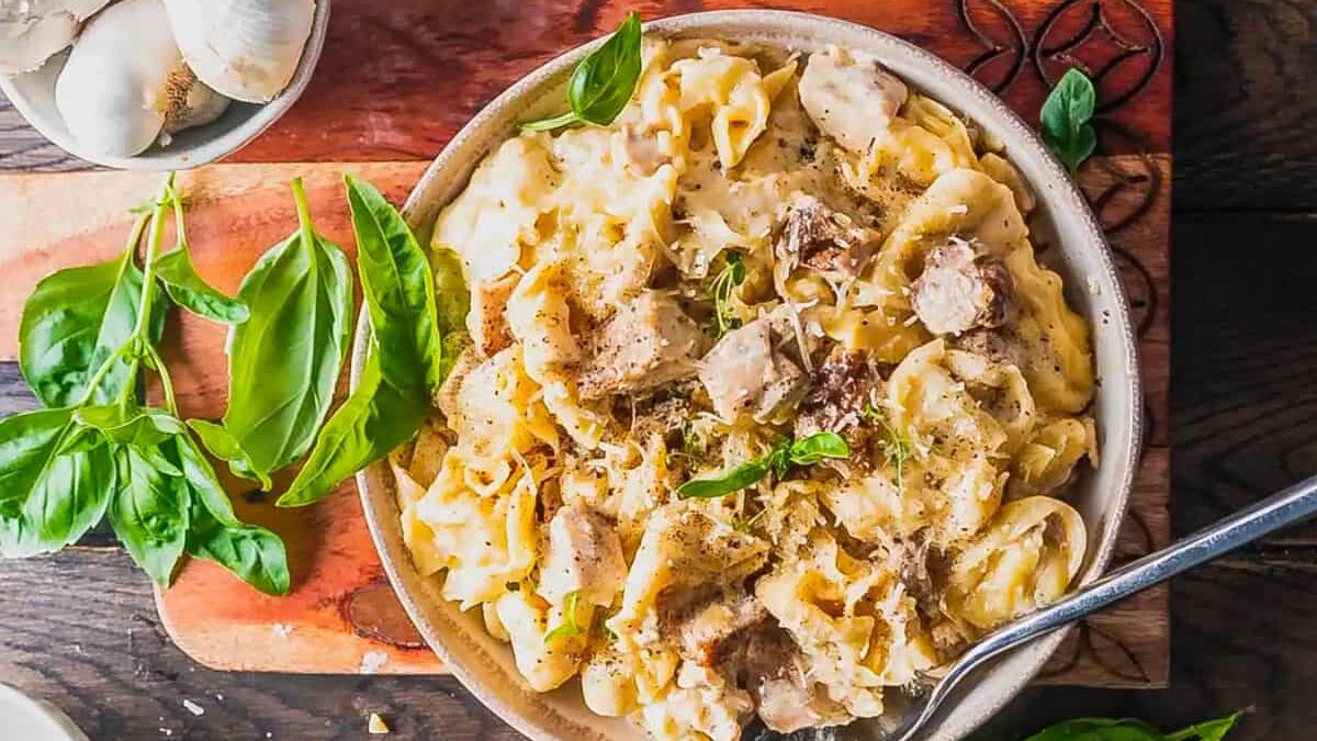 A bowl of creamy chicken and mushroom pasta garnished with fresh basil leaves sits on a wooden board. Whole garlic bulbs and additional basil leaves are placed nearby. A fork rests in the bowl.