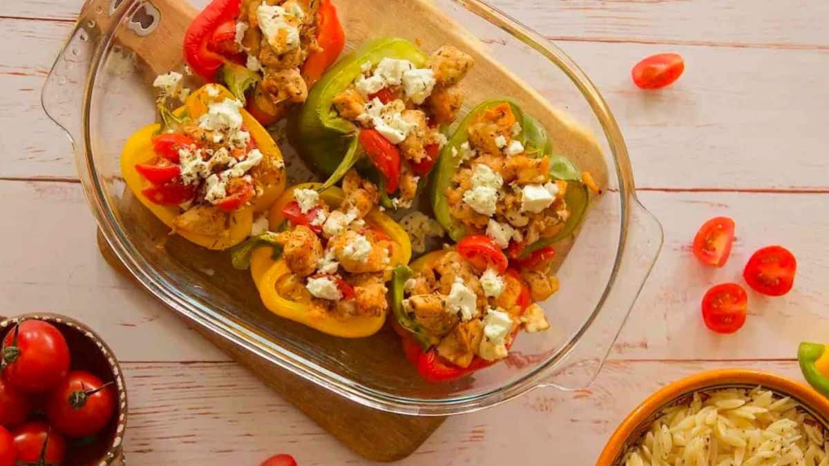 A glass baking dish with colorful stuffed bell peppers filled with chicken, cherry tomatoes, and feta cheese on a wooden board. Nearby are a bowl of cherry tomatoes and a bowl of cooked orzo pasta.
