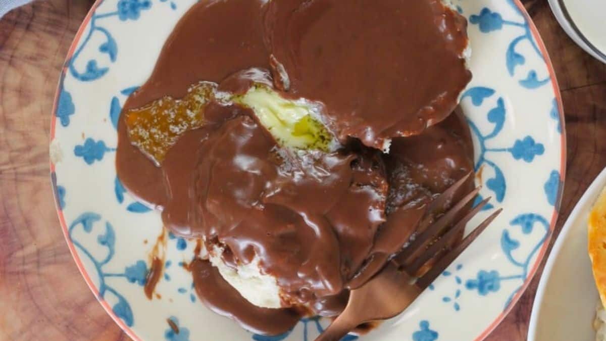 A ceramic plate with a blue and white pattern holds a biscuit covered in thick chocolate gravy. A fork is placed on the plate's edge. The dish is set on a wooden table.