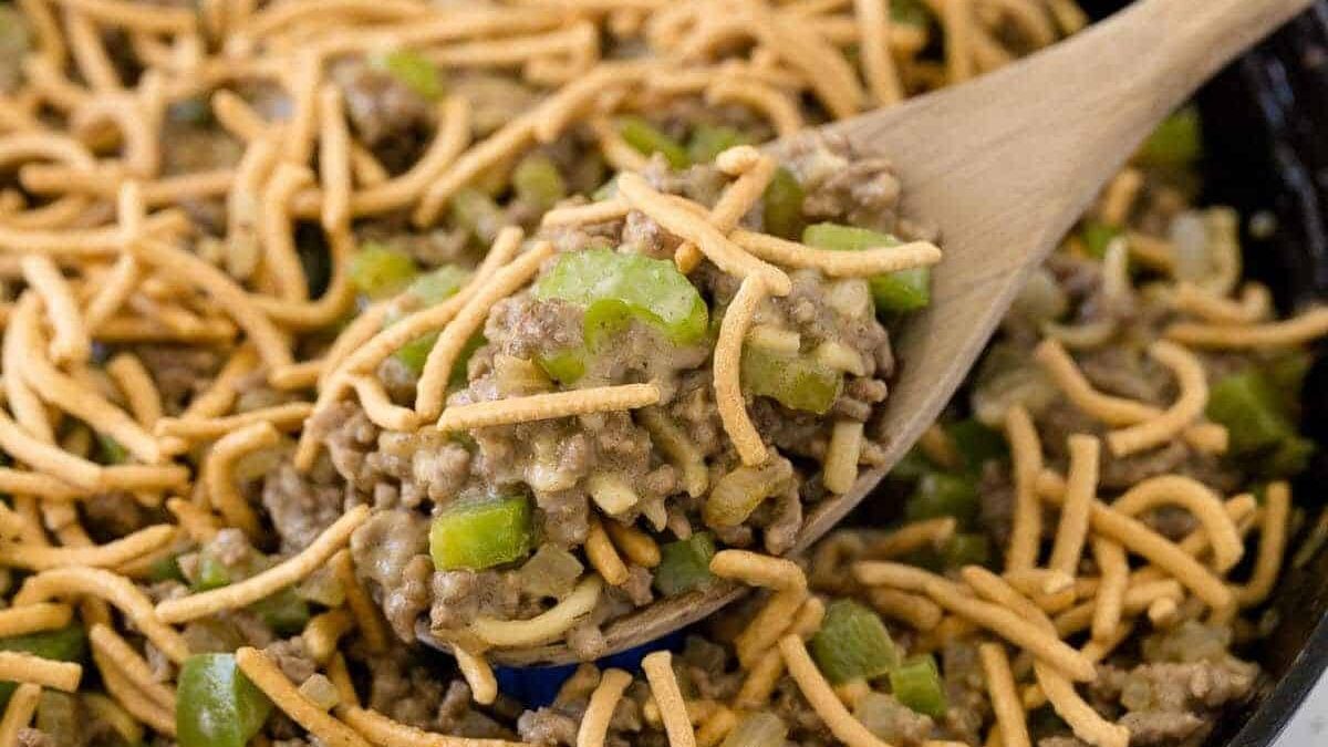 A wooden spoon scoops a serving of a dish made with ground beef, chopped green bell peppers, and crispy chow mein noodles. The mixture is in a skillet, showcasing a hearty and textured appearance.