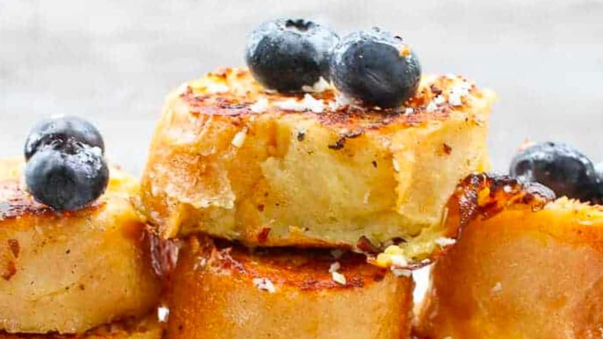 Stack of golden-brown French toast rounds topped with fresh blueberries. The toast is crispy on the edges, and some pieces are drizzled with a hint of syrup, creating a delicious and inviting look.