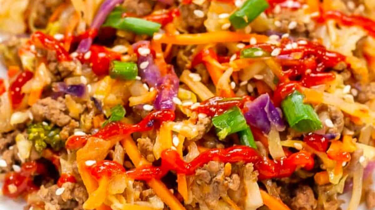 Close-up of a colorful stir-fry dish featuring ground meat, shredded carrots, chopped green onions, purple cabbage, and broccoli, all drizzled with a vibrant red sauce and sprinkled with sesame seeds. The ingredients look fresh and appetizing.