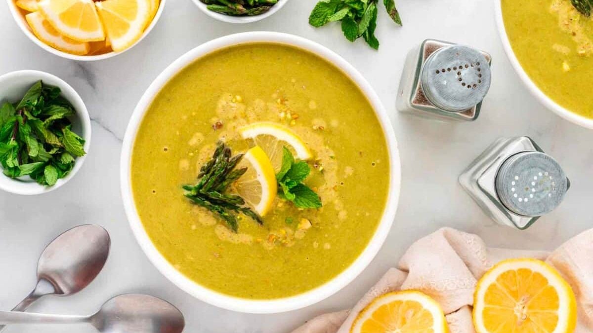 A bowl of green vegetable soup garnished with lemon slices and asparagus sits on a marble surface. Surrounding it are bowls of lemon wedges, fresh herbs, and a bundle of asparagus. Two spoons, a napkin, and salt and pepper shakers are also visible.