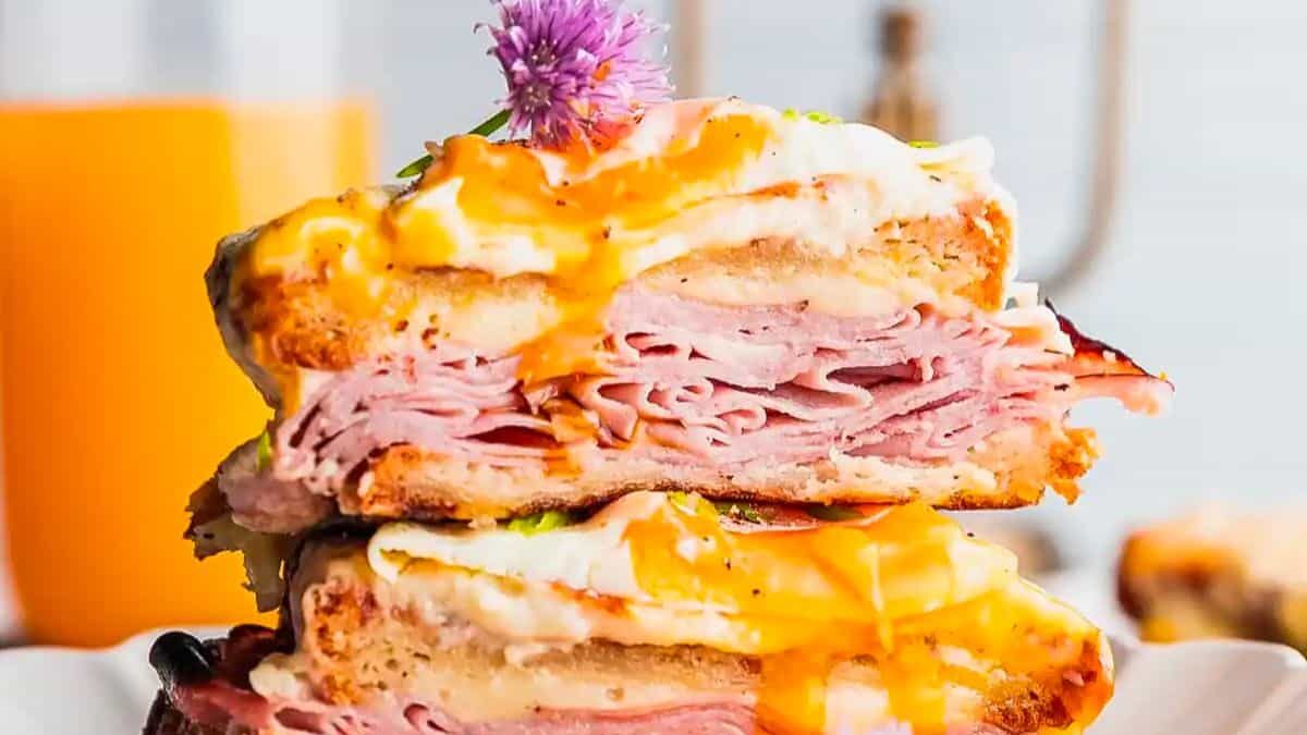 A close-up of a stacked sandwich showcasing layers of thinly sliced ham, melted cheese, and a fried egg with runny yolk. The sandwich is garnished with a purple flower and served on a white plate, with an orange drink in the background.