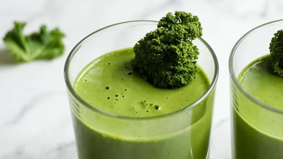 Two glasses filled with a green smoothie, topped with a sprig of curly kale. A small piece of kale is in the background on a white marble surface.
