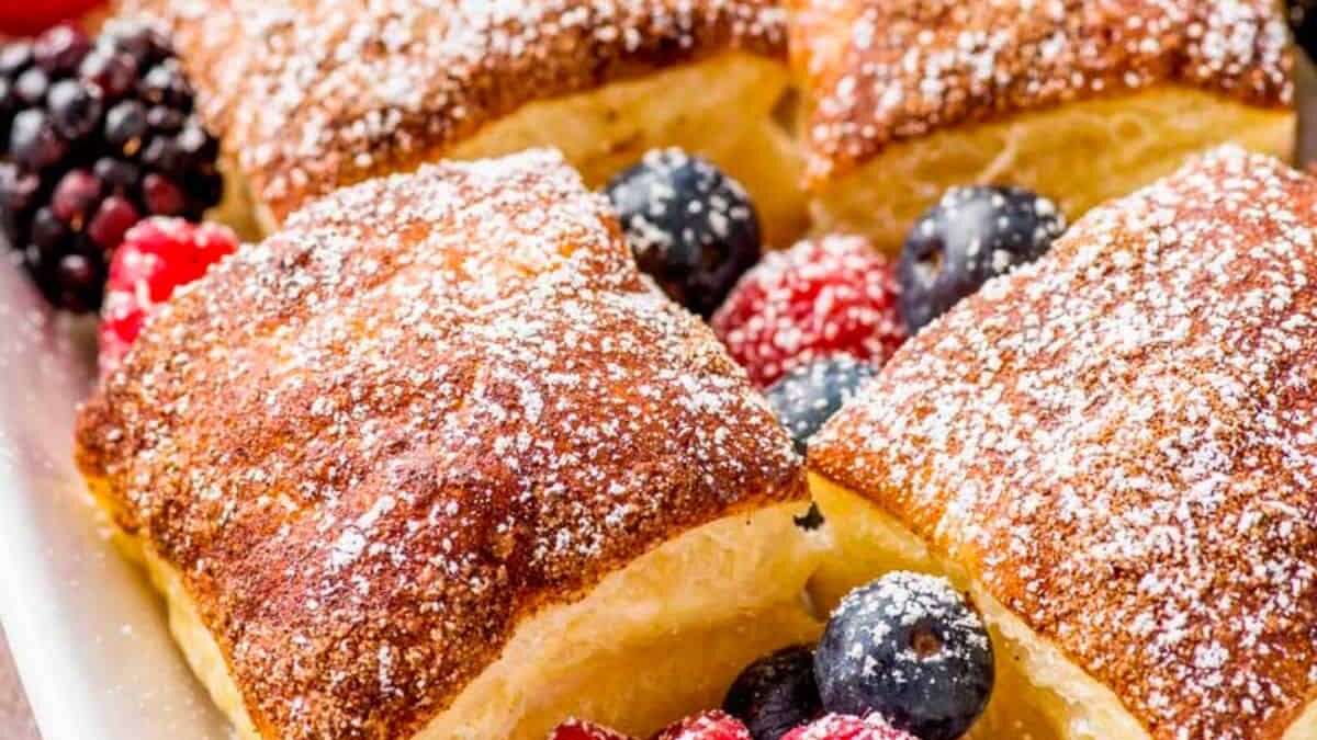 Golden brown pastry squares dusted with powdered sugar are surrounded by fresh mixed berries, including blueberries, raspberries, and blackberries, creating a colorful and appetizing dessert display.