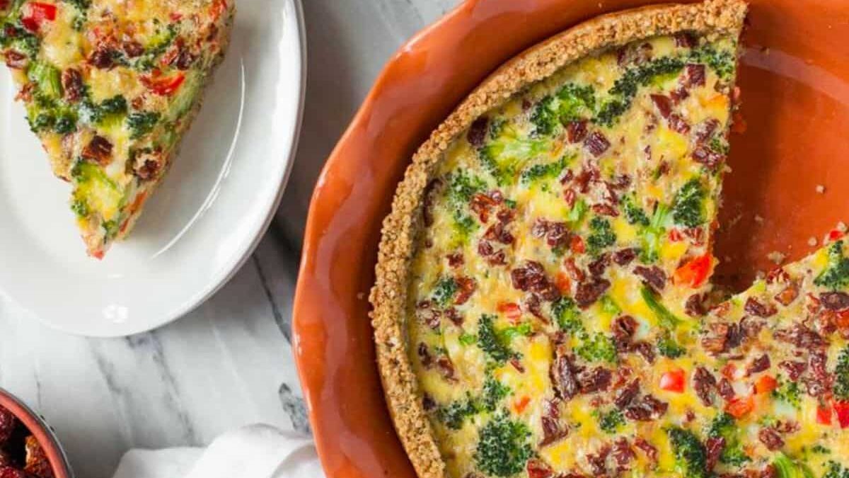A slice of vegetable quiche with broccoli, red bell peppers, and bacon sits on a white plate next to the main dish in a round, brown pie dish. The quiche has a golden crust and is on a marble surface.