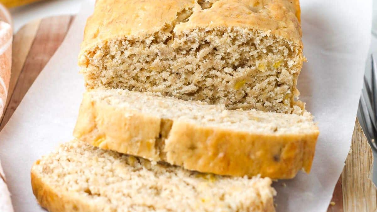 A loaf of banana bread on parchment paper, with three slices cut in front. The bread has a golden-brown crust and a moist interior with visible banana pieces. It rests on a wood surface.