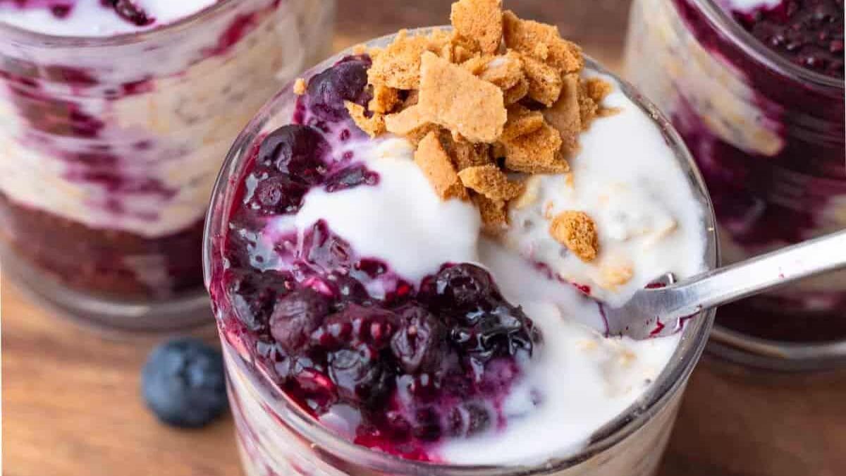 Glass jar filled with layers of creamy yogurt, oats, and dark berry compote, topped with crumbled graham crackers. A spoon is resting inside the jar, and other similar jars are partially visible in the background.