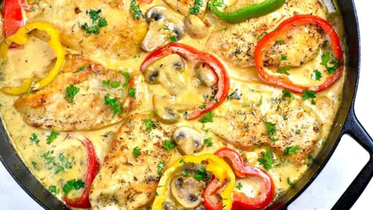 Chicken breasts cooked in a creamy sauce with sliced mushrooms, red, yellow, and green bell peppers, and garnished with fresh parsley. The dish is presented in a black skillet.