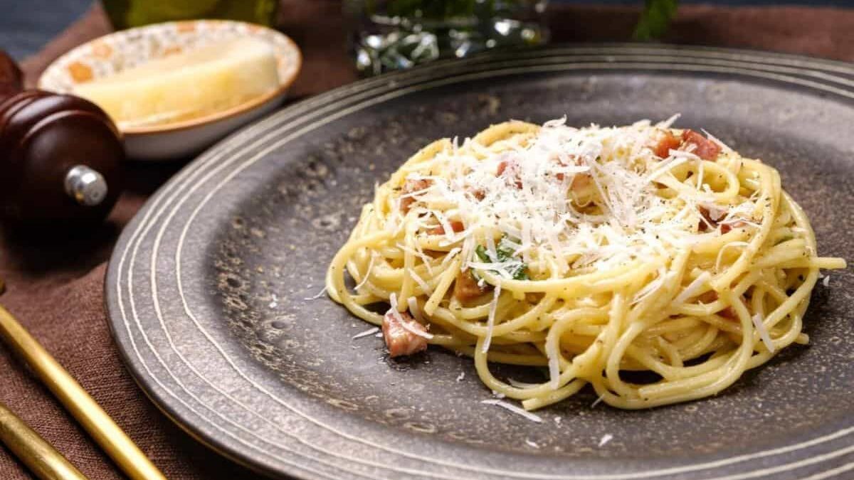 A plate of spaghetti carbonara topped with grated cheese and pieces of ham, on a dark, textured plate. There is a pepper grinder, silverware, cheese, olive oil, and a plant in the background.