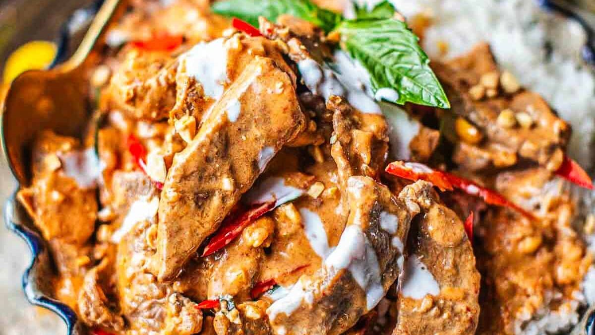 A close-up of a spicy Thai beef dish with creamy red curry sauce, garnished with sliced red chilies and fresh basil leaves, served over a bed of rice.