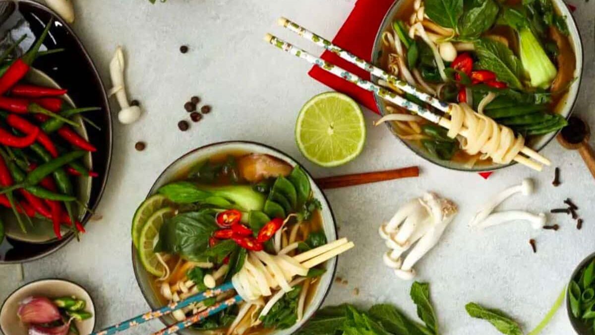 Two bowls of noodle soup garnished with greens, red chilies, lime slices, and mushrooms on a light surface. Surrounding items include fresh herbs, chilies, mint, garlic, and spices. Chopsticks rest on the bowls, and a red napkin is nearby.