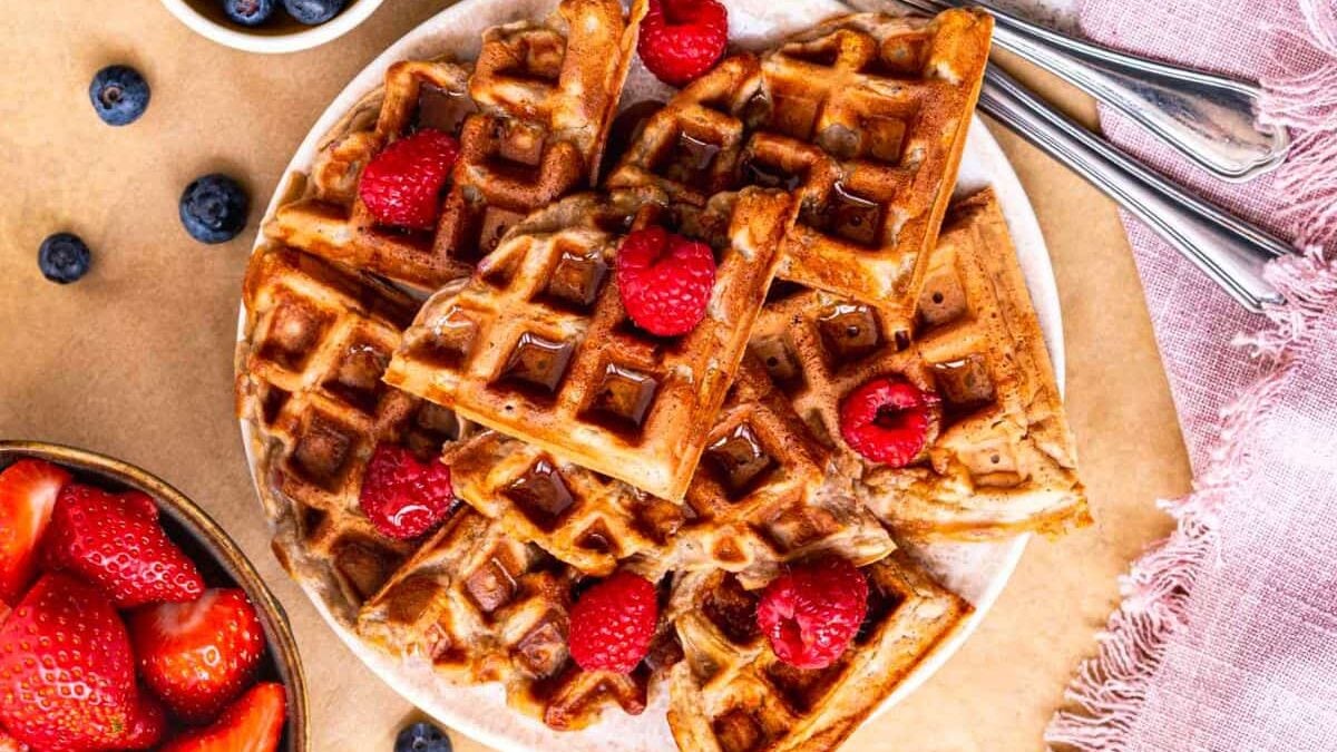 A plate of golden brown waffles topped with fresh raspberries. A bowl of strawberries and blueberries is nearby. Two forks rest on a pink textured cloth beside the dish.