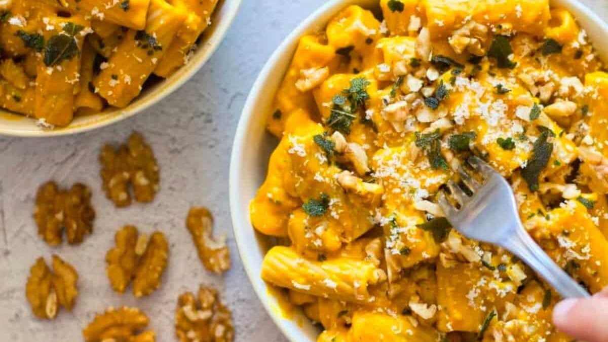 Two bowls of creamy pasta with a rich orange sauce are garnished with chopped walnuts, crumbled cheese, and herbs. A fork is twirling pasta from one bowl, while several shelled walnuts are scattered on the white surface nearby.