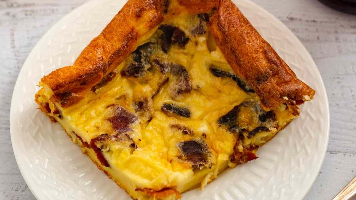 A slice of Yorkshire pudding filled with a savory mixture is served on a white plate. The pudding is golden brown with visible chunks of meat and vegetables, offering a delicious combination of textures and flavors.