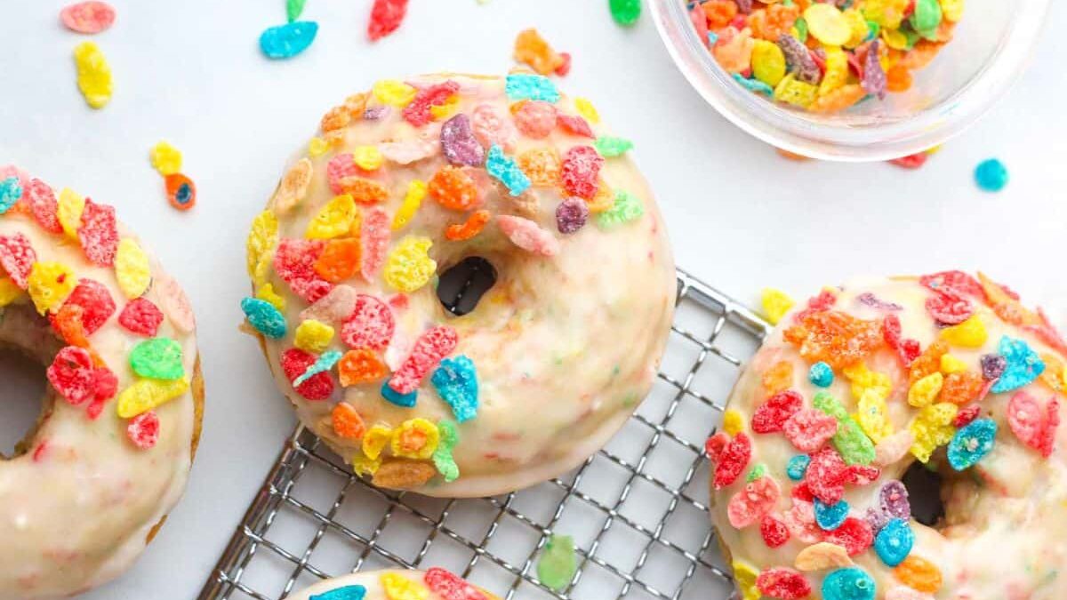 Four glazed donuts topped with colorful cereal pieces are displayed on a wire cooling rack. A small bowl filled with more cereal pieces sits nearby, with some of the cereal scattered around on a white surface.