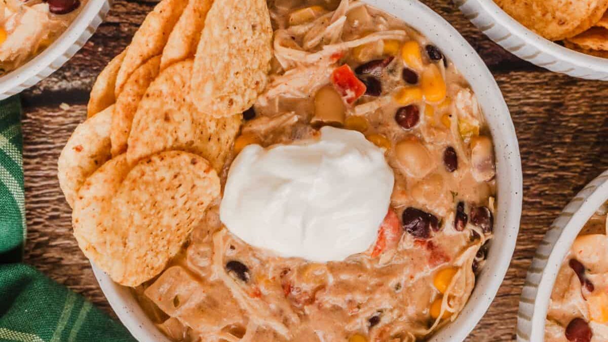 A bowl of creamy chicken taco soup topped with a dollop of sour cream and garnished with tortilla chips. The soup contains corn, black beans, and chunks of chicken, and is served on a rustic wooden surface with part of a green cloth visible.