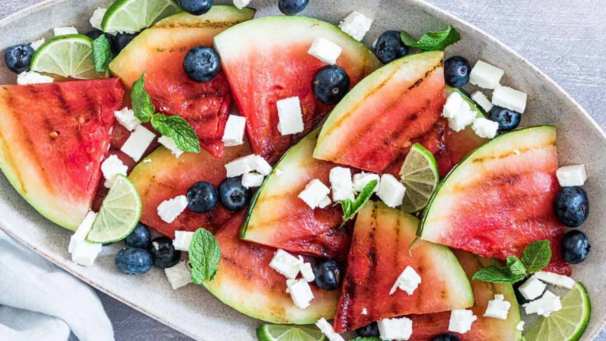 A platter of grilled watermelon wedges topped with blueberries, feta cheese cubes, mint leaves, and lime slices. The colorful summer salad is arranged in an oval dish on a light grey surface.