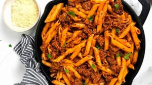 A skillet filled with penne pasta mixed with a rich, meaty tomato sauce, garnished with fresh parsley. A small bowl of grated cheese and a striped napkin are placed beside the skillet on a white surface.