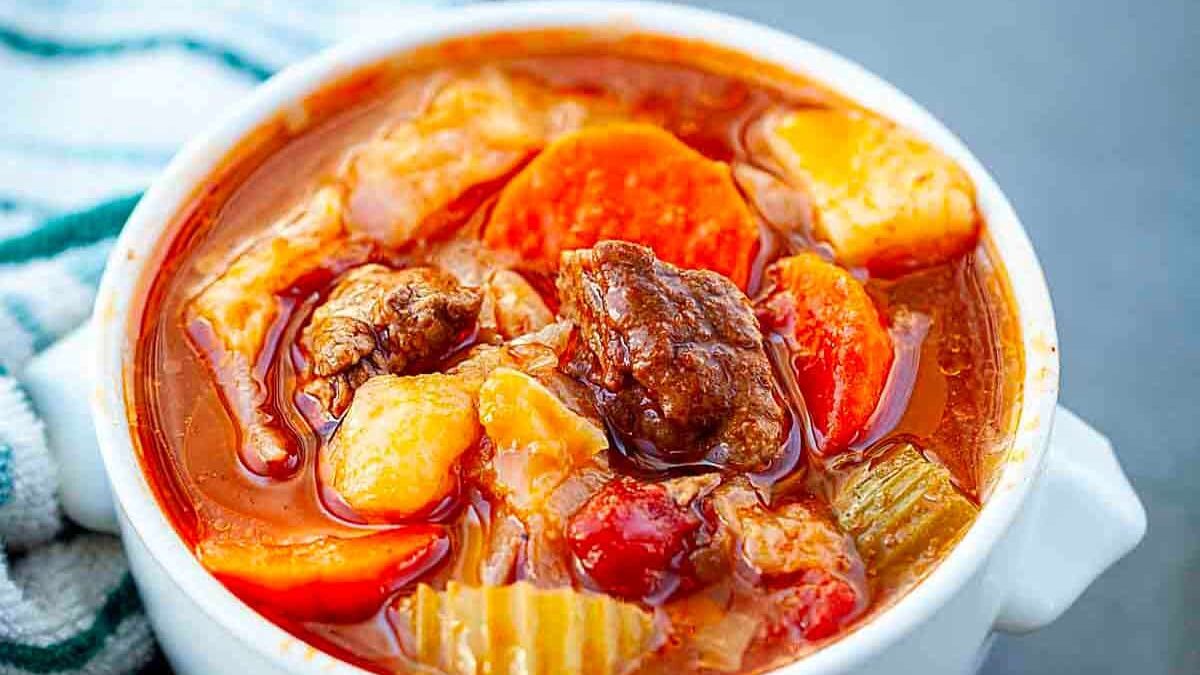 A bowl of hearty beef stew filled with chunks of beef, carrots, potatoes, celery, and tomatoes in a rich broth. The bowl is white and sits on a gray surface next to a striped cloth.