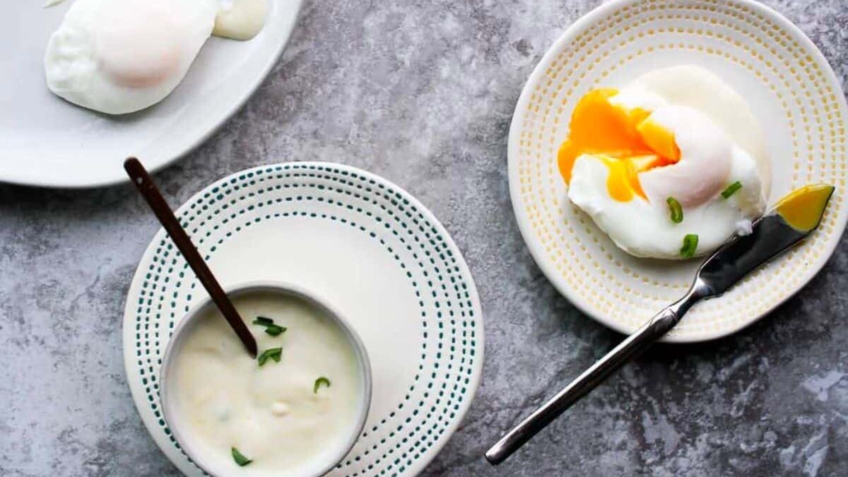 A grey marble surface with two plates each holding poached eggs. One egg is intact, while the other is cut open with a runny yolk. A small bowl of creamy sauce garnished with green herbs sits nearby, with a knife resting on one plate.