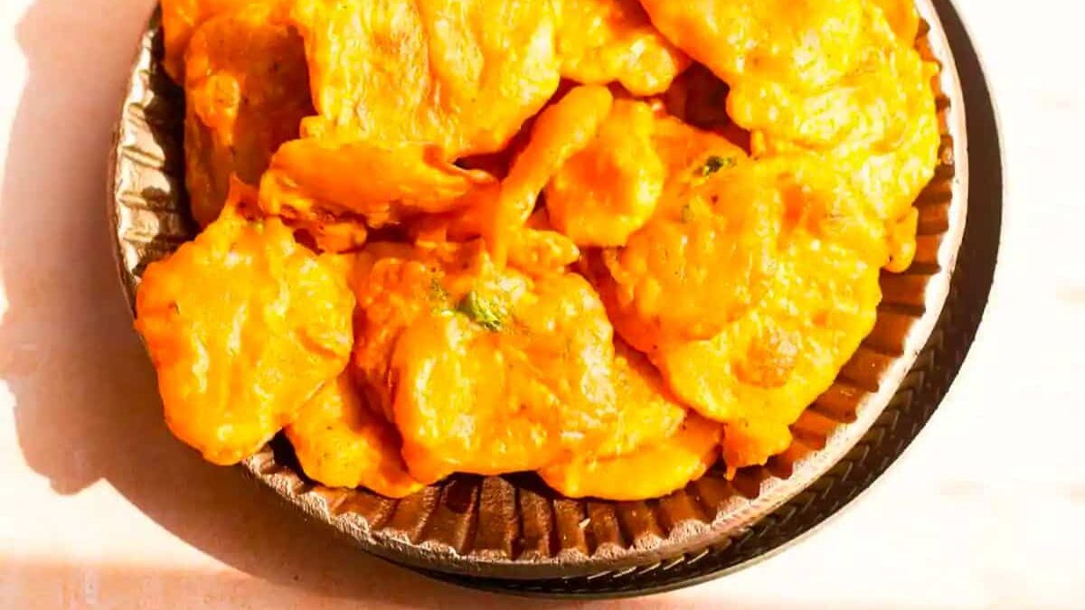 A plate filled with vibrant orange fritters, possibly Indian pakoras, served on a brown, round dish. The fritters appear crispy and are piled on each other, filling the plate.