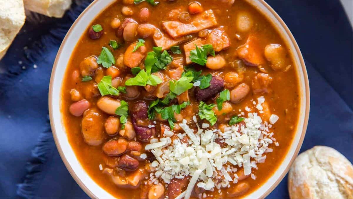A bowl of hearty bean stew garnished with chopped parsley and grated cheese. The stew contains a variety of beans and pieces of vegetables in a rich, thick sauce. Pieces of bread are visible on the side, and the bowl rests on a dark blue cloth.