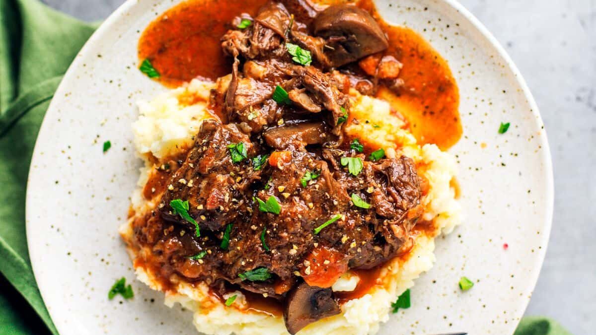 A plate of mashed potatoes topped with savory beef stew and mushrooms, garnished with chopped parsley. A fork rests beside the dish on a speckled, round plate, with a green cloth partially visible to the side.