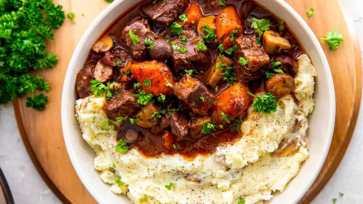 A hearty bowl of beef stew with chunks of beef, carrots, and mushrooms in a rich sauce, served alongside creamy mashed potatoes. Garnished with fresh parsley, all presented on a wooden board.