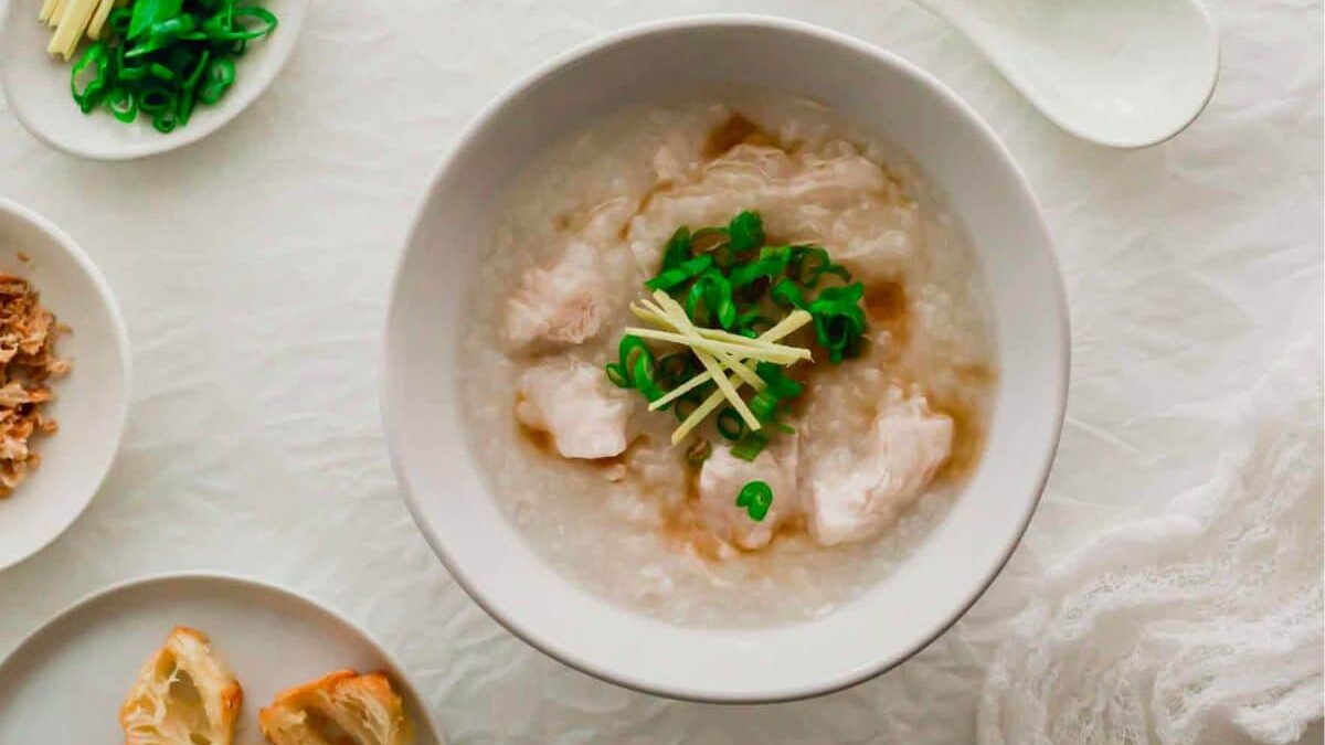 A white bowl filled with rice porridge, garnished with green onions and sliced ginger sits on a white textured surface. Side dishes including chopped green onions, fried dough pieces, and shredded ginger are placed around the bowl. White spoons are next to the bowl.