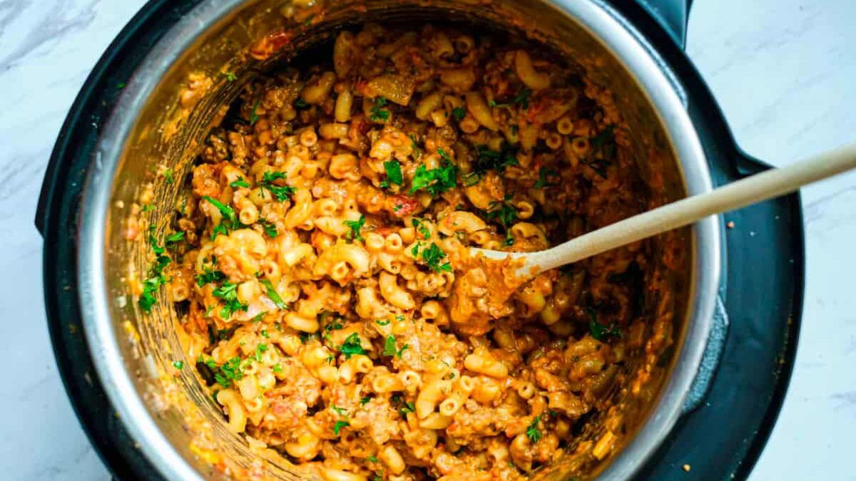 An overhead view of a pressure cooker filled with cooked macaroni pasta mixed with ground meat, sauce, and garnished with fresh herbs. A wooden spoon is resting inside the cooker, ready to stir.