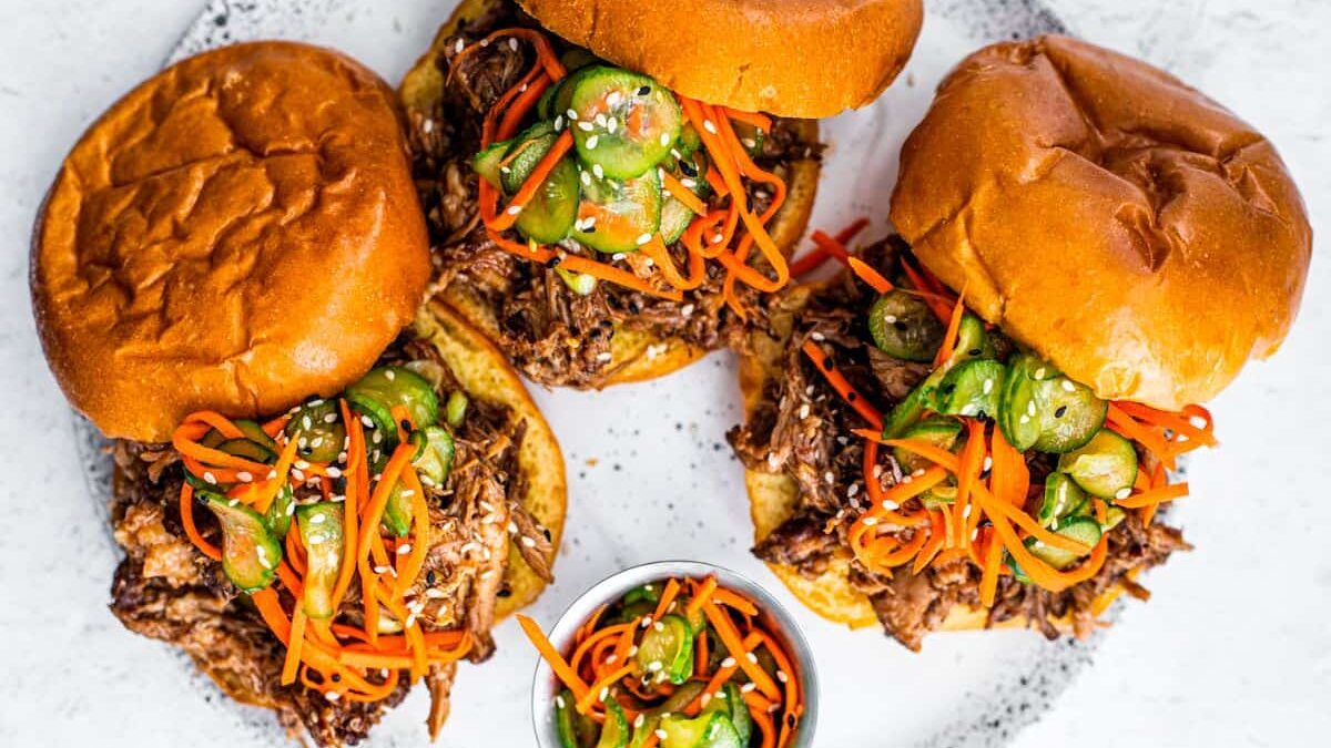 Three pulled pork sandwiches topped with pickled vegetables on a white speckled plate, accompanied by a small bowl of extra pickled veggies, are set against a white background. The buns are golden and slightly toasted.
