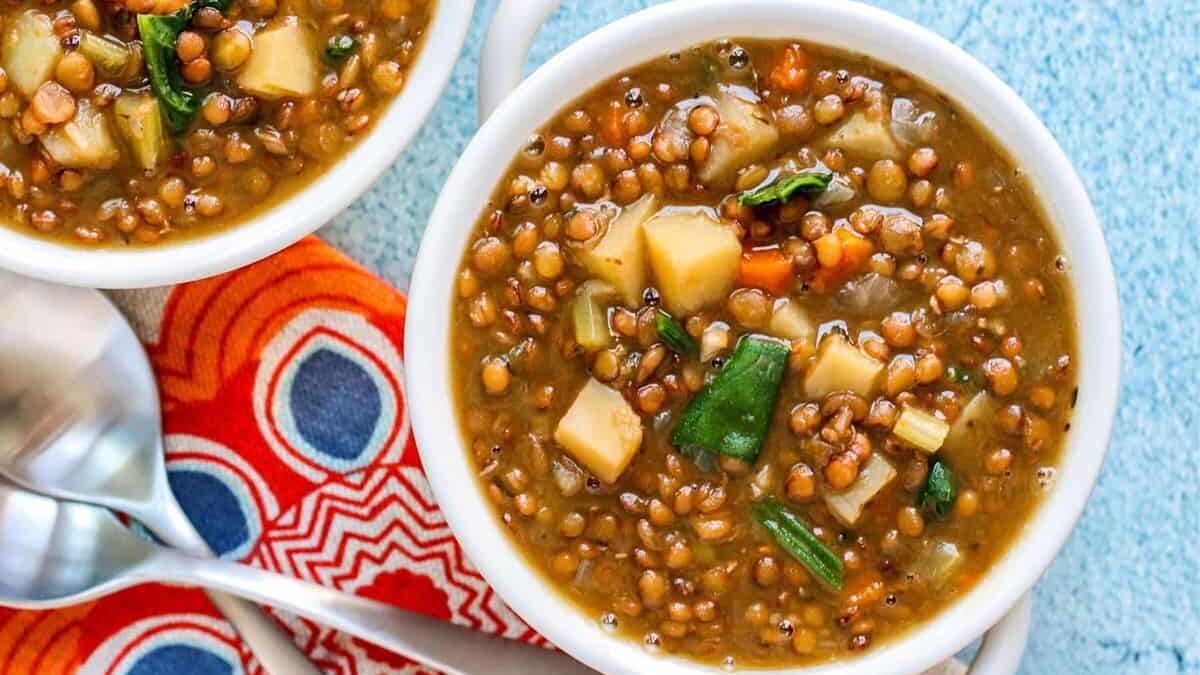 Two bowls of lentil soup with diced vegetables, including carrots and spinach, are placed on a blue surface. The bowls are accompanied by a colorful red and orange patterned napkin and two silver spoons.